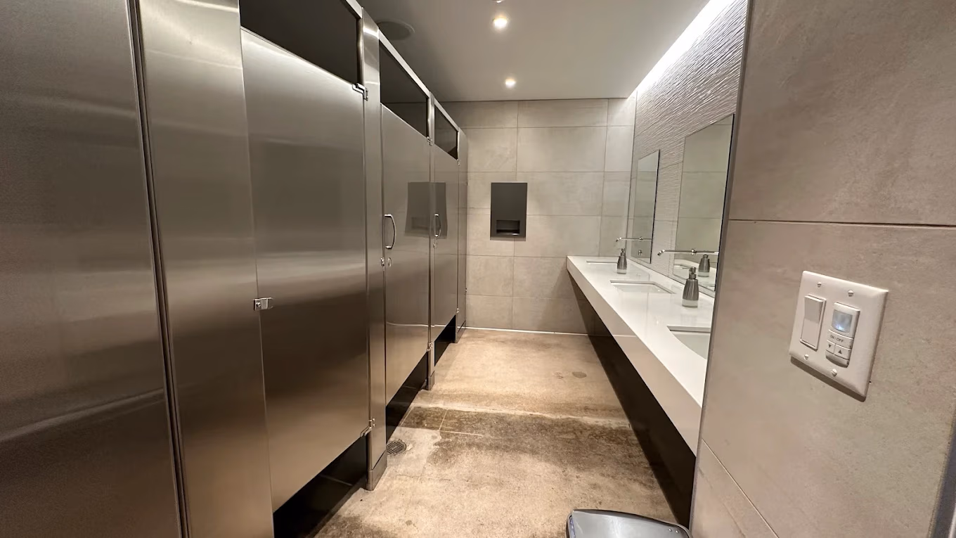 Modern public restroom with stainless steel stalls, white sinks, mirrors, and tiled walls and floor.