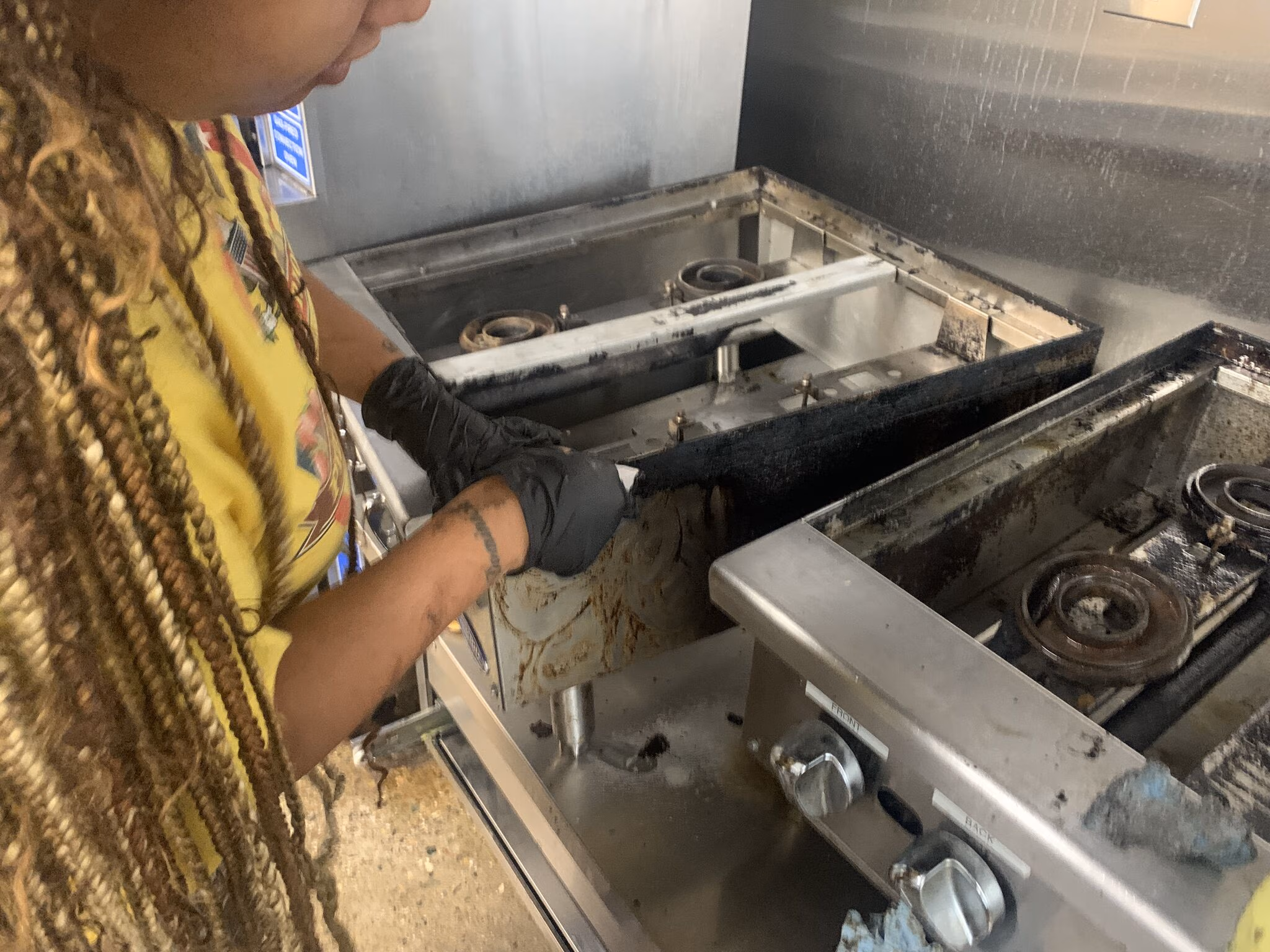 Person wearing black gloves cleaning a greasy stove burner in a kitchen.