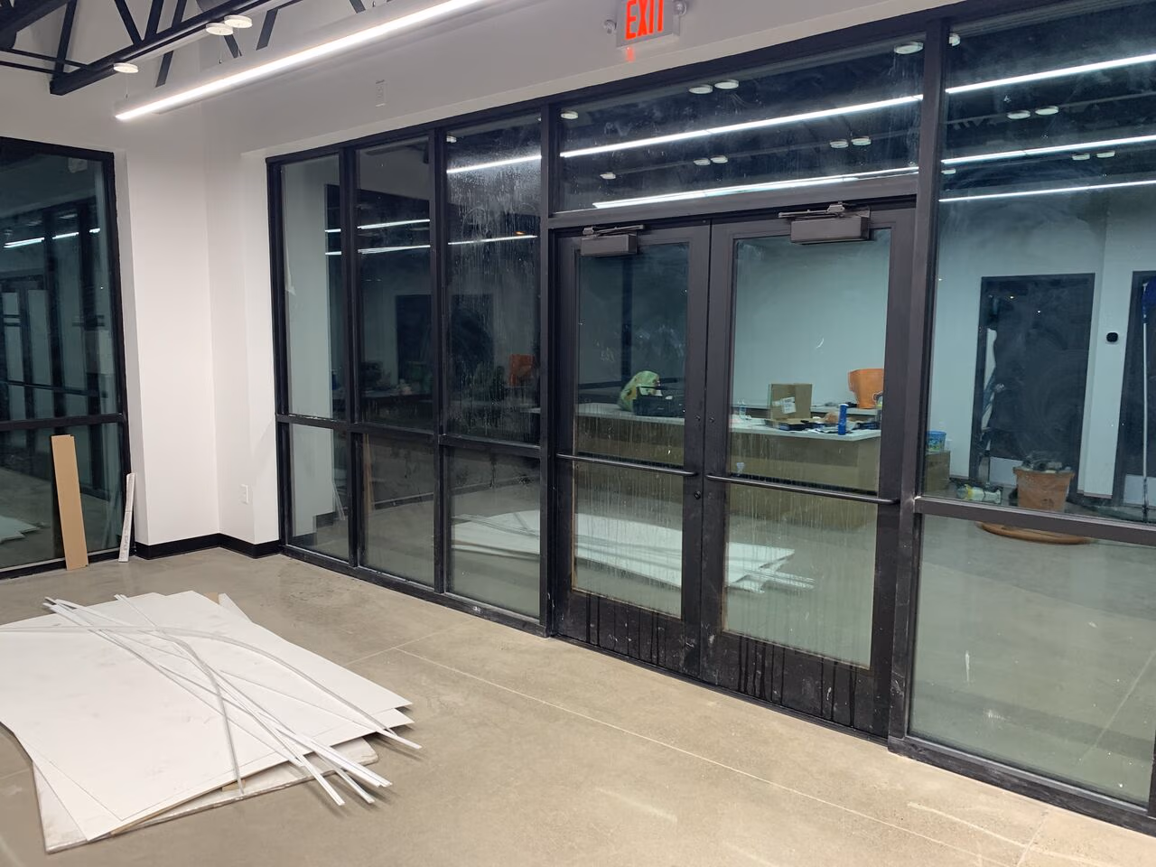 Indoor space with glass doors, scattered construction materials, and an illuminated exit sign above.