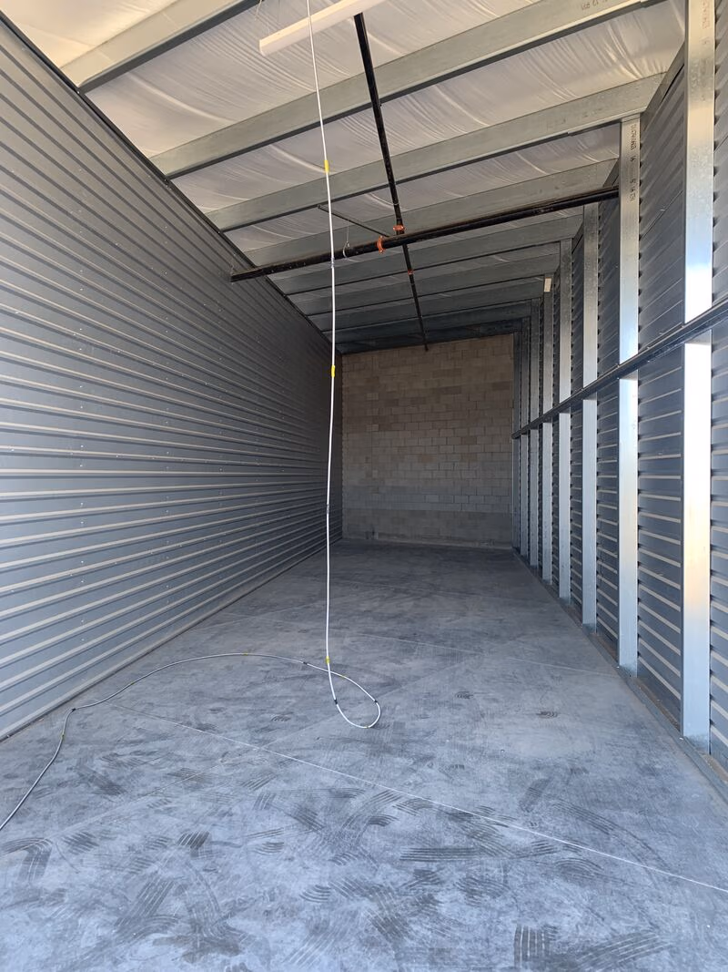 Empty industrial storage room with metal walls, concrete floor, ceiling beams, and a hanging white cable.