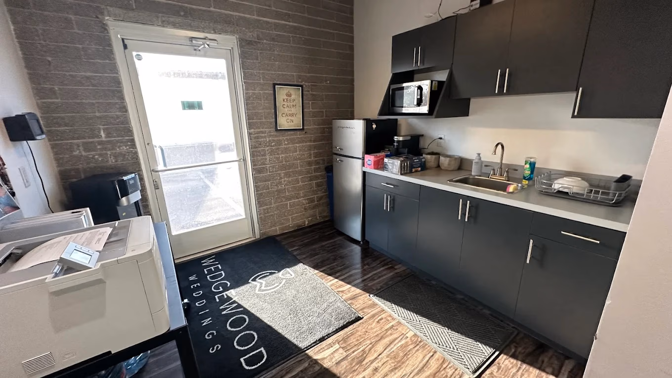 Small office kitchenette with refrigerator, microwave, sink, and printer near an exit door with a Wedgewood Weddings mat.