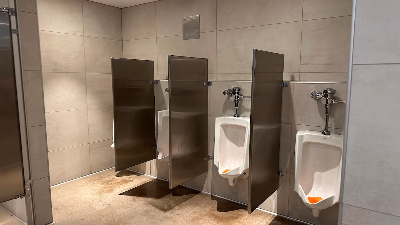 Three white urinals with stainless steel privacy dividers in a public restroom with beige tiled walls.