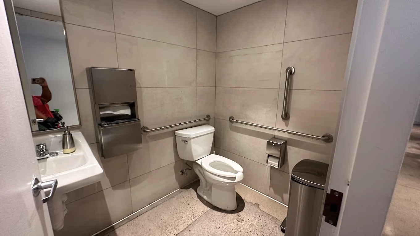 Accessible public restroom with white toilet, grab bars, sink with soap dispenser, and trash can.