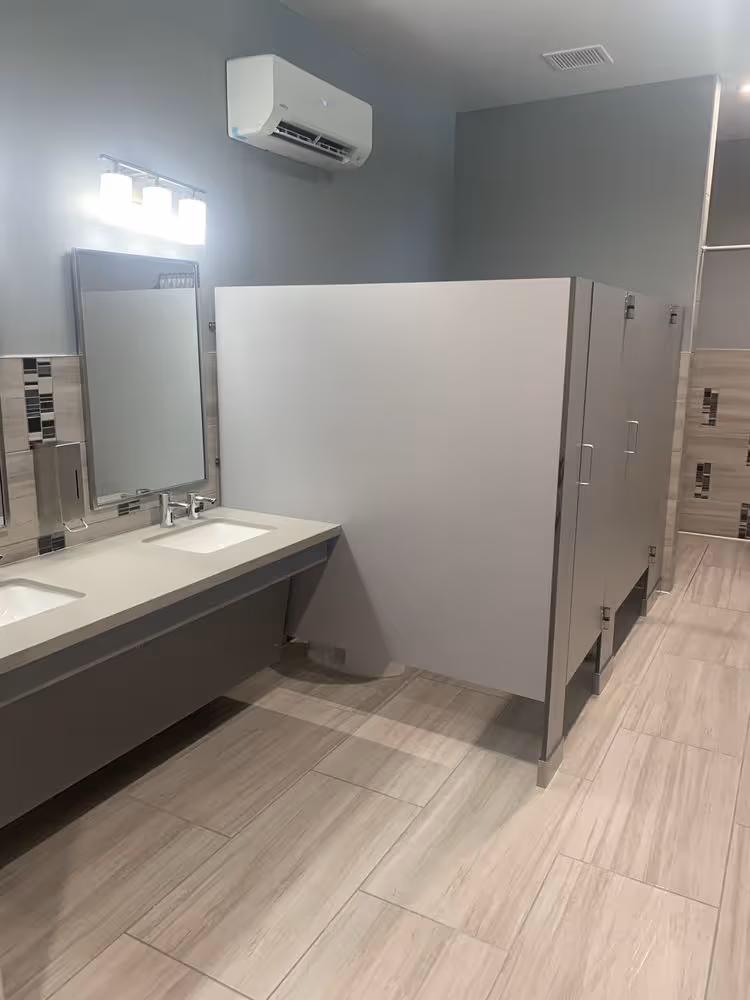 Clean public restroom with two sinks, a mirror, three closed toilet stalls, and an air conditioning unit.