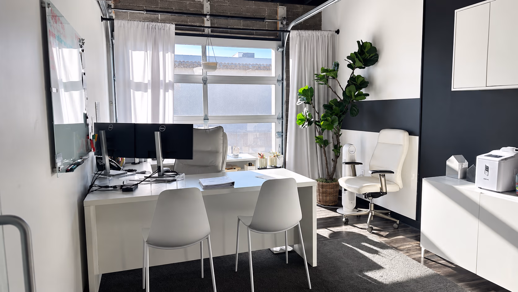 Modern office with white desks, two monitors, white chairs, a plant, and large window with curtains.