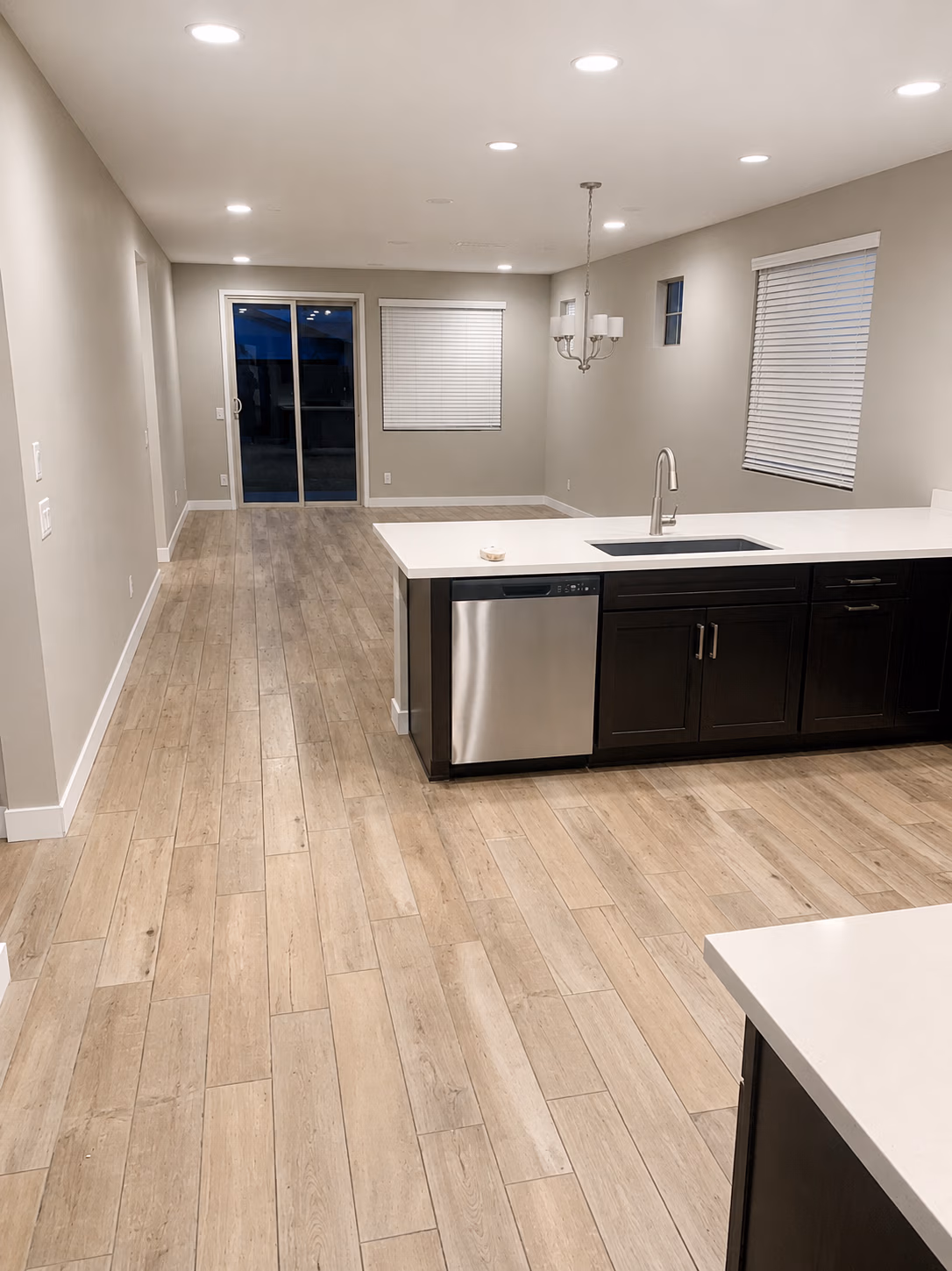 Empty modern kitchen and dining area with wood-look tile floor, white countertops, and dark cabinets.