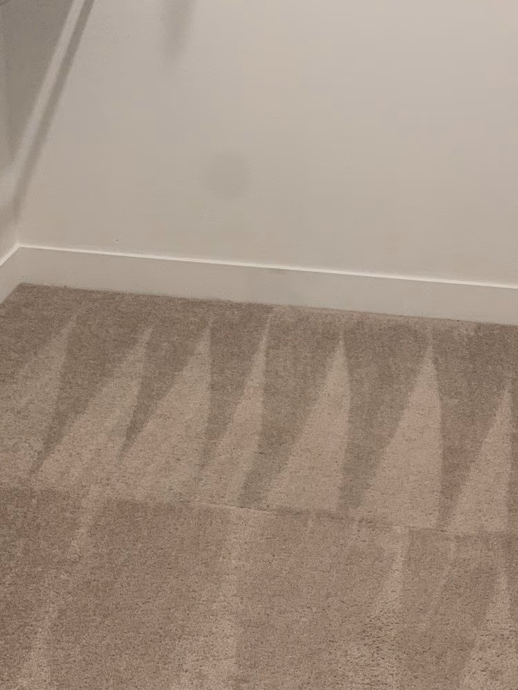 Light brown carpet with triangular vacuum cleaning patterns next to a plain white wall and baseboard.