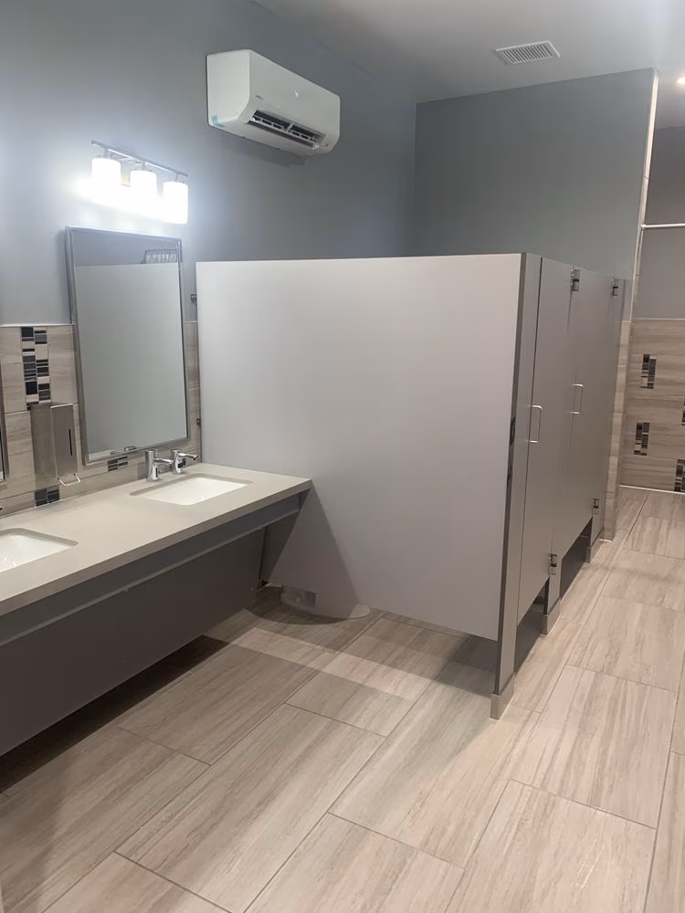 Clean public restroom with two sinks, a mirror, three closed toilet stalls, and an air conditioning unit.