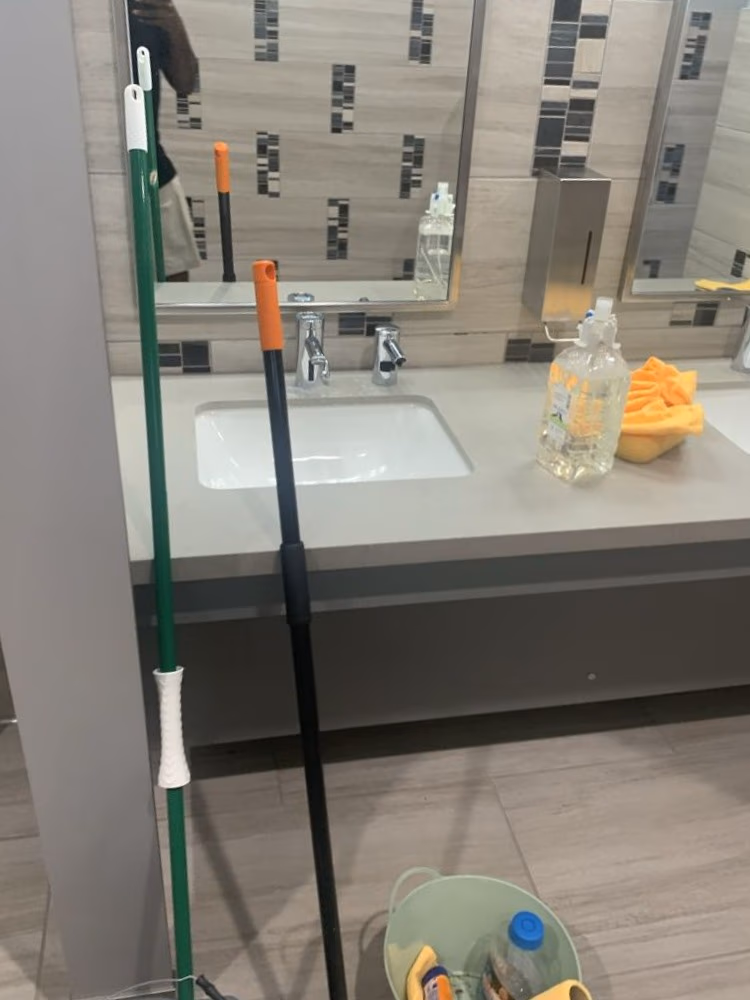 Cleaning supplies including mop, bucket with sponges and bottle, next to a sink in a tiled restroom.