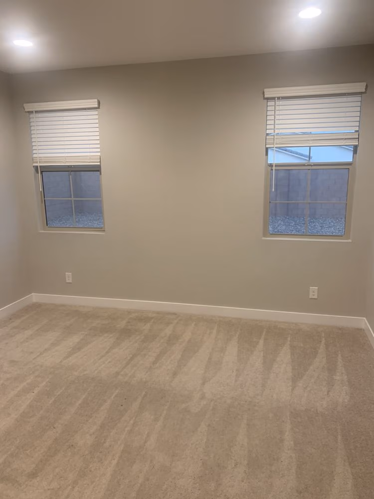 Empty beige carpeted room with two windows covered by white blinds and recessed ceiling lights.