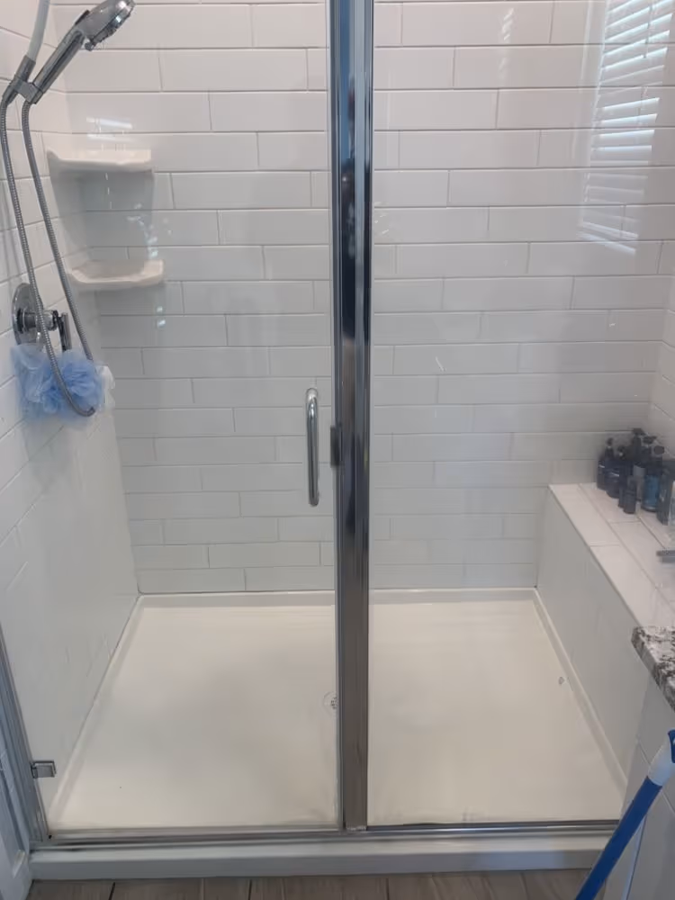 White tiled walk-in shower with glass door, handheld showerhead, blue loofah, and bench with bottles.