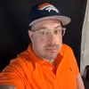 Man wearing glasses, orange polo, and Denver Broncos cap, standing against a dark background.