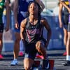 Athlete in black kneeling on track ready for a sprint race, with other runners nearby.