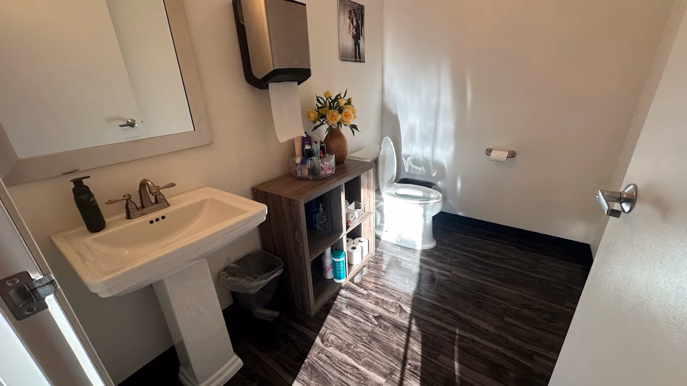 Bathroom with pedestal sink, mirror, open toilet, wooden shelf with toiletries and flowers, dark wood floor.