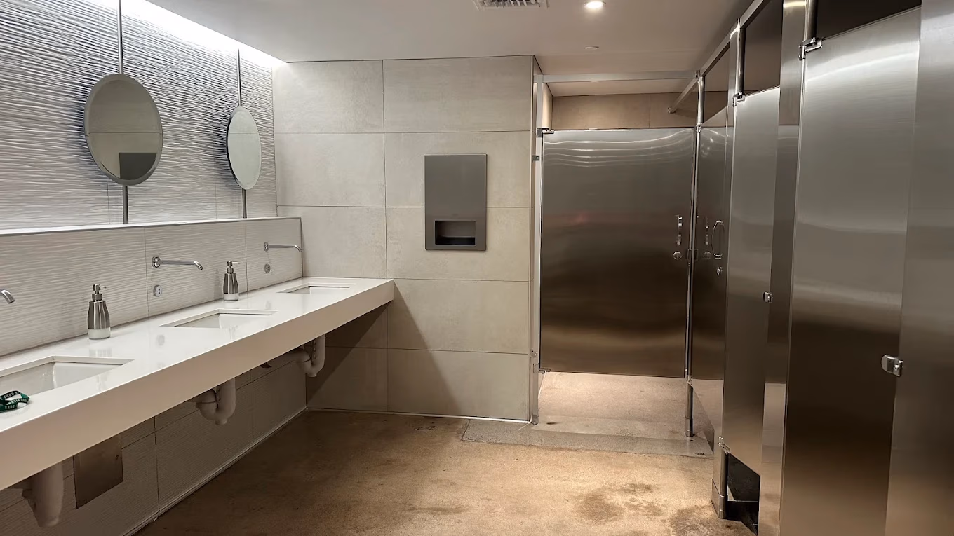 Modern public restroom with three sinks, round mirrors, soap dispensers, and stainless steel toilet stalls.