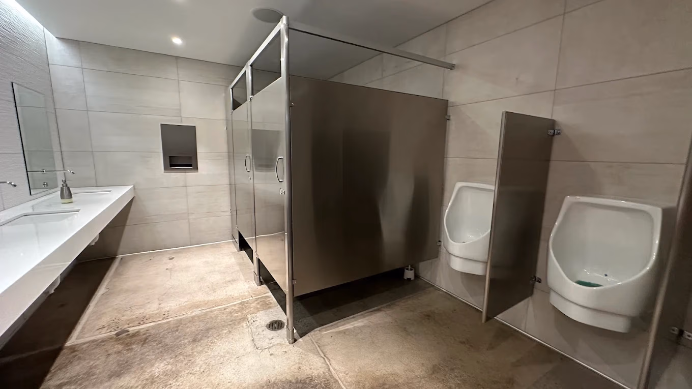 Modern public restroom with stainless steel stalls, two urinals with privacy dividers, and a white sink counter.