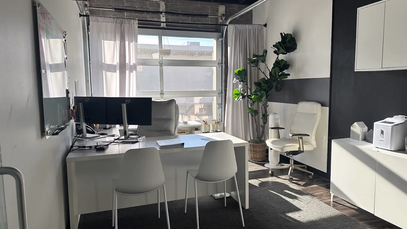 Bright modern office with white furniture, dual monitors, chairs, potted plant, and window with curtains.